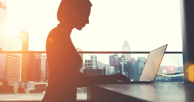 Photo of a silhouette of a woman working ona laptop in front of a city skyline.